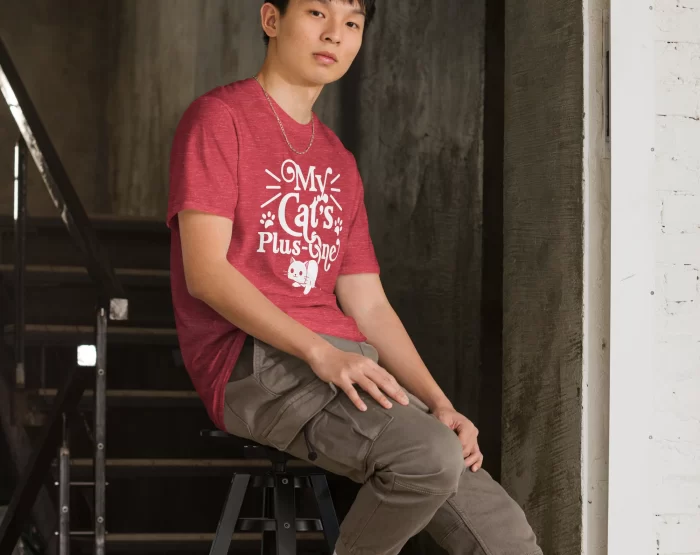 Young man sitting on stairs wearing heather red "My Cat’s Plus One" shirt in urban setting