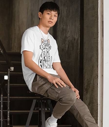 Young man wearing a white “Proud Spare Human” t-shirt from Spare Human Society, sitting on steps inside with a relaxed expression.