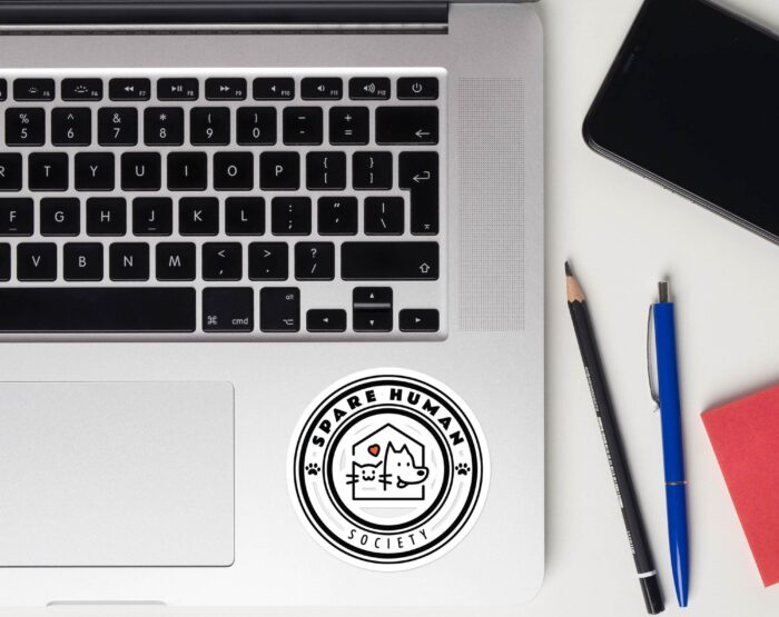 funny-pet-parent-laptop-decal-black-and-white. Spare Human Society sticker placed on silver laptop next to keyboard and pens