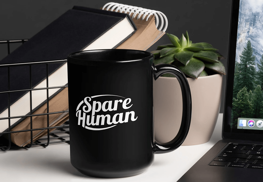 Black coffee mug with “Spare Human” printed on it, sitting on a desk with notebooks, a laptop, and a succulent — the true fuel station for every certified Spare Human.