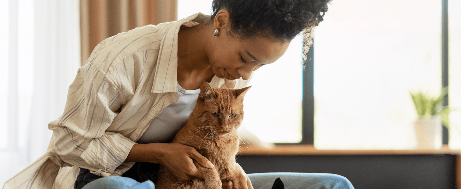 Smiling woman gently cuddling her orange tabby cat indoors, capturing the cozy, judgment-free bond between pets and their humans.
